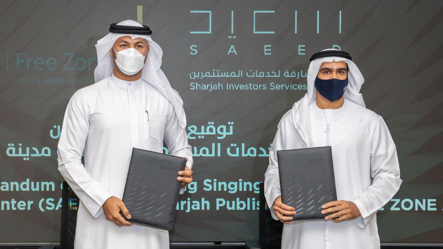 Two officials in traditional attire hold documents, standing in front of SPC Free Zone and SAIF Zone logos.
