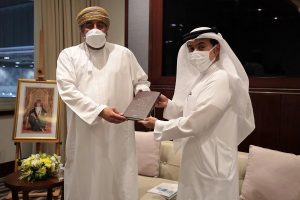 Two men exchanging a document in an office setting with a portrait in the background.