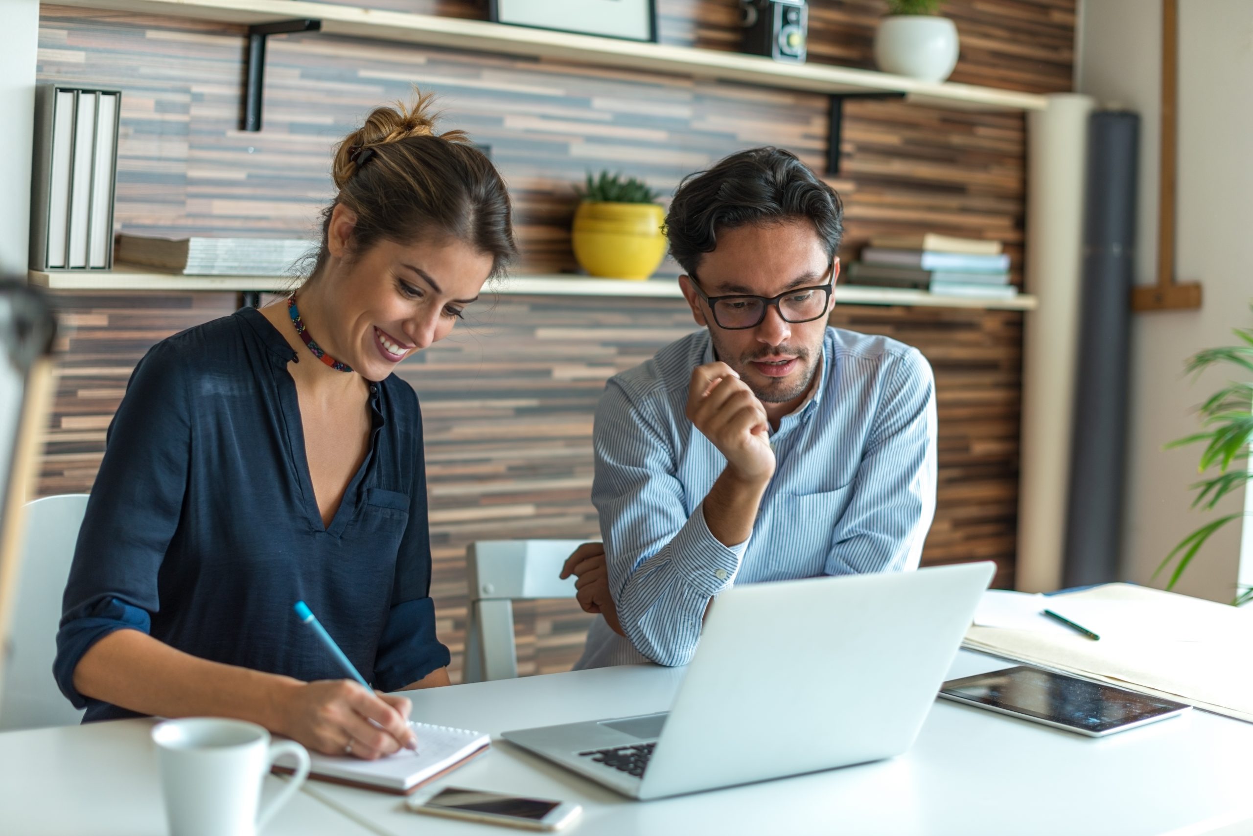 A man and woman discuss business at a laptop with a notebook and coffee cup.