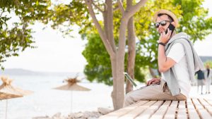 Man with laptop on beach, talking on phone, wearing yellow hat and sunglasses.